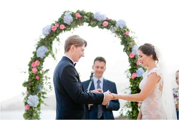 The couple is exchanging rings during an outdoor wedding ceremony, with the groom in a navy suit and black bow tie placing a ring on the bride's finger while she smiles downward. The bride wears a lace cap-sleeve gown with a veil. An officiant in a navy suit stands behind them, smiling. The backdrop is a large circular floral arch decorated with lush greenery, pink roses, and blue hydrangeas. The shot is a medium portrait framing the couple and officiant from approximately the waist up.