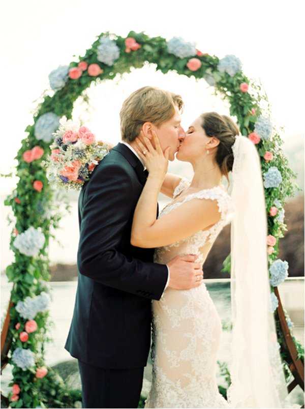 A couple shares their first kiss at an outdoor ceremony in front of a circular floral arch constructed from lush greenery, coral/pink roses, and powder blue hydrangeas. The groom wears a navy suit, while the bride wears a fitted lace gown with an open back and a long cathedral-length veil with her hair up. The bride holds a bouquet of coral roses, blue hydrangeas, pink blooms, and white wildflowers. The portrait-style shot is slightly soft in focus with a bright, airy exposure, and the arch rests on a visible wooden A-frame stand.