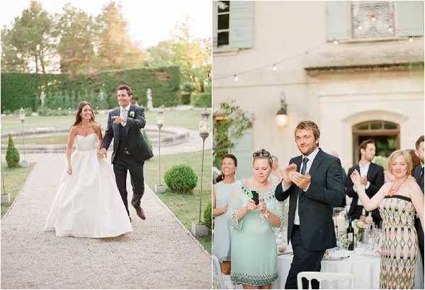 A diptych of two wedding photographs taken at a French chateau estate during golden hour. On the left, the bride and groom walk joyfully along a gravel garden path, with the bride wearing a strapless white A-line gown and the groom in a dark navy suit with a white boutonniere; the formal garden behind them features topiary, a reflecting pool, and stone statues. On the right, wedding guests applaud and react during what appears to be a cocktail hour or outdoor reception on the chateau's terrace, with the stone facade of the chateau and mint-green shuttered windows visible in the background; string lights are strung overhead, and guests are dressed in smart-casual attire including a mint green dress and a patterned strapless dress. Both images have a soft, warm film-style edit with a light and airy aesthetic. Potential venue feature image.