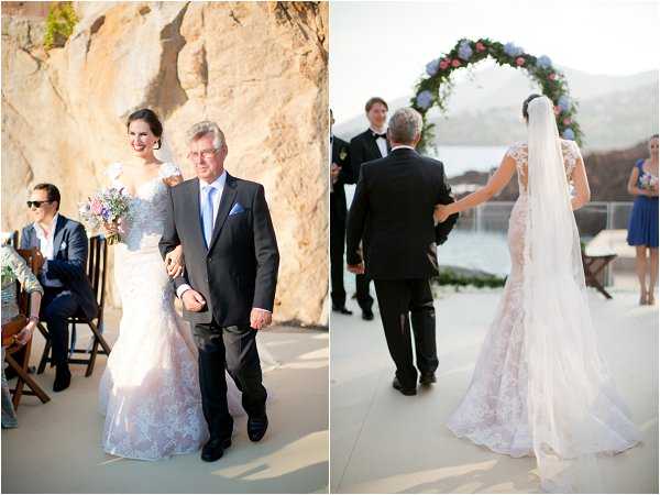 Two side-by-side images capturing the processional moment of an outdoor wedding ceremony on a coastal terrace. On the left, the bride walks arm-in-arm with an older man, likely her father, both facing the camera; she wears a lace gown with a blush/ivory tone and carries a bouquet of mixed purple and white blooms, while he wears a dark suit with a light blue tie. On the right, the same pair is photographed from behind as they approach the groom and officiant, with the bride's cathedral-length veil and lace train trailing behind her; a circular floral arch decorated with pink, white, and blue flowers frames the altar area in the background. Seated guests in dark and navy attire are visible on either side of the aisle. The setting is an open-air terrace with a rocky cliff backdrop and water visible in the distance, suggesting a Mediterranean coastal venue. Both images are medium-wide ceremony shots with natural daylight.