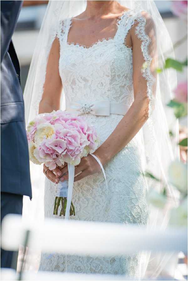 A close-up portrait of a bride during what appears to be an outdoor ceremony, framed from the shoulders to the knees. She wears a fitted white lace dress with wide lace-trimmed straps, a square neckline, and a satin bow belt accented with a rhinestone brooch, paired with a lace-edged cathedral veil. She holds a compact round bouquet of soft pink hydrangeas and cream peonies tied with a white satin ribbon. A figure in a dark suit is partially visible to her left, and blurred floral arrangements in pink tones are visible in the background.