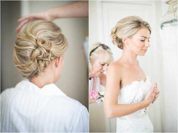 A two-image panel from the bride's getting-ready preparations, shot indoors in a bright, light-filled room. The left image is a close-up portrait from behind showing the bride's blonde hair styled in a twisted and pinned updo with soft waves, while a stylist's hands are visible at the top of the frame. The right image shows the bride in profile, eyes closed, wearing a strapless white lace wedding dress with a scalloped neckline, while an older woman — likely a family member — fastens the back of the dress; the assistant is wearing a pink rose corsage on her wrist. Both shots are soft and well-lit with a pale, neutral interior background and a dress hanging on a hanger visible in the right frame. The overall styling is classic and polished.