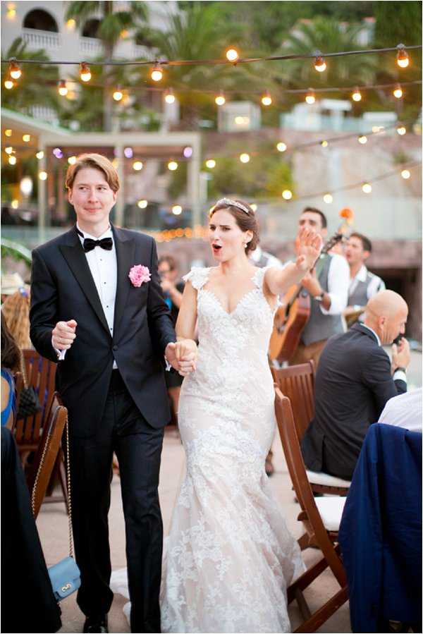 The bride and groom make their grand entrance at an outdoor evening reception, walking hand-in-hand through seated guests with expressions of excitement and joy. The bride wears a fitted lace gown with cap sleeves, a V-neckline, and a crystal headband, while the groom wears a classic black tuxedo with a black bow tie and a pink floral boutonniere. The reception is set on an open-air terrace with strings of warm Edison bulb bistro lights strung overhead, creating a festive atmosphere, and a live musician playing guitar is visible in the background. The setting appears to be a resort or villa courtyard, and the overall decor style is classic with warm, relaxed lighting.