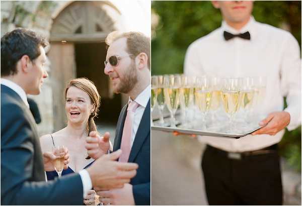 A diptych of two cocktail hour scenes shot outdoors. On the left, three guests converse animatedly in front of a stone building entrance; a woman in a navy dress holds a champagne flute and laughs, flanked by two men in navy suits — one wearing sunglasses and a blush pink tie. On the right, a close-up portrait of a waiter in a white dress shirt and black bow tie holding a silver tray lined with approximately 15 filled champagne flutes. Both images are warm-toned and suggest a classic, formal wedding reception style with a champagne service element.