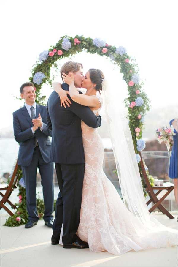 The bride and groom share their first kiss during an outdoor ceremony on a terrace or elevated platform, with water visible in the background. They stand beneath a circular floral arch densely covered in greenery, blue hydrangeas, and pink roses. The bride wears a blush lace fitted gown with a long cathedral veil, while the groom is dressed in a dark navy suit. An officiant or guest in a navy suit stands to the left applauding, and a bridesmaid in a cobalt blue dress is partially visible to the right. The decor palette combines navy, blush, and periwinkle blue in a classic coastal style. Medium-distance portrait shot.