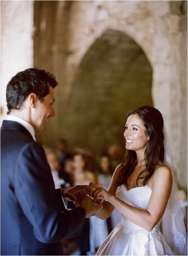 The bride and groom are exchanging rings during an indoor ceremony set within a stone chapel or ancient church, with a rounded stone archway visible in the background. The groom wears a navy suit with a white dress shirt, while the bride wears a strapless white ballgown with lace detailing and a sheer veil. The bride has long dark wavy hair worn half-up and is smiling directly at the groom as he places the ring on her finger. A blurred group of seated guests is visible in the background. The image is a mid-length portrait shot with soft, warm natural light giving it a film photography quality.
