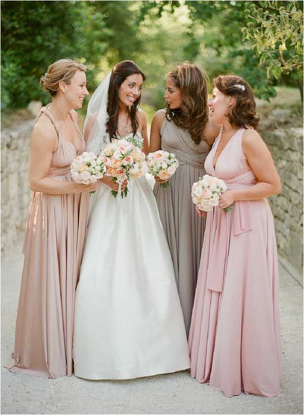 A bridal party portrait taken outdoors, showing a bride with three bridesmaids standing together and conversing. The bride wears a white satin ball gown with a cathedral-length veil and carries a bouquet of peach and ivory roses with greenery. The three bridesmaids wear floor-length multiway wrap dresses in mismatched but coordinating neutral tones — blush pink, taupe-grey, and dusty rose — each carrying a matching bouquet of cream and soft peach roses. The styling has a soft, neutral palette with a classic, understated aesthetic. The shot is a medium portrait taken at eye level with a low stone wall and blurred green foliage visible in the background.