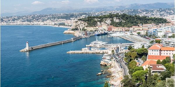 This image does not depict a wedding photograph. It is an aerial wide shot of a Mediterranean coastal city, showing a harbor with a jetty, lighthouse, marina with moored boats, and urban architecture extending up a hillside. No people, wedding events, or wedding-related elements are visible. The image appears to be a cityscape or venue location overview shot, likely of Nice, France. Potential venue feature image.