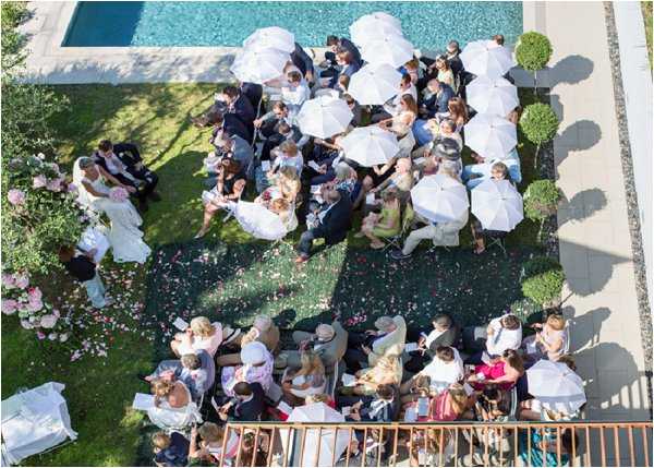 An aerial wide shot of an outdoor wedding ceremony taking place on a lawn beside a rectangular swimming pool. Approximately 60-80 guests are seated in chairs arranged in rows, many shading themselves with white parasols due to bright sunlight. The bride in a white gown and groom in a dark suit stand at the altar on the left side of the frame, accompanied by what appears to be an officiant and bridal party members. The aisle is scattered with pink and white flower petals leading up to the ceremony area, which is flanked by arrangements of pink roses. Neatly trimmed topiary balls in planters line the right side of the space, and a section of additional guests are seated on a terrace in the lower portion of the frame. The overall decor palette is white and soft pink with a classic, garden-party aesthetic.