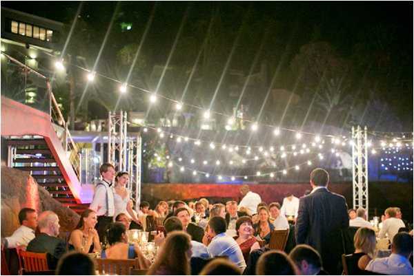 An outdoor evening wedding reception with approximately 50-60 guests seated at round tables covered with white linens and lit by candles. The setting appears to be a terrace or rooftop venue with urban buildings visible in the background, strung with warm-toned globe string lights suspended between metal truss poles overhead. A person, likely a speaker or emcee, stands at the right foreground facing the crowd, while the bride in a white dress and a man in a white shirt and dark suspenders are visible standing near a staircase with red railings on the left side. The decor style is relaxed and informal, with wooden chairs and candlelit tables. Wide-angle shot capturing the full reception scene at night.
