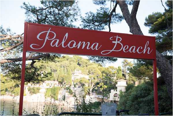 Close-up shot of a red metal entrance sign reading 'Paloma Beach' in white cursive lettering, mounted on two red posts. The background shows a coastal setting with pine trees, Mediterranean-style architecture, and water visible in the distance. No people are present in the frame. Potential venue feature image.