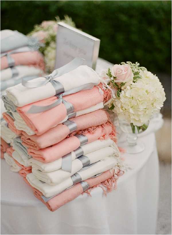 Close-up detail shot of a guest favor or comfort station table at an outdoor wedding reception. Two stacks of folded pashmina wraps in alternating coral/salmon and ivory are tied with silver satin ribbon bows and arranged on a white linen-covered table. A third smaller stack of light blue-grey pashminas is partially visible on the left. A small floral arrangement of cream hydrangeas and blush roses in a clear glass vase sits to the right of the stacks, and a framed sign is propped in the background. The decor palette is coral, ivory, and silver-grey, consistent with a classic and polished wedding styling theme.
