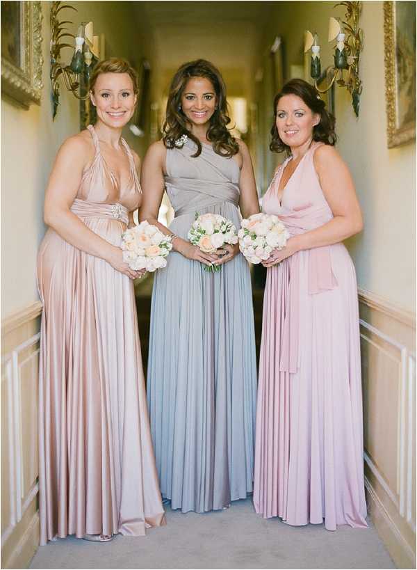 Three bridesmaids pose together in a portrait shot taken indoors in a carpeted hallway, likely within a chateau or manor house, with ornate gold-framed wall sconces and paneled wainscoting visible along the corridor. Each bridesmaid wears a floor-length multiway wrap dress in a different soft pastel shade: the left wears blush nude, the center wears dove grey-blue, and the right wears dusty mauve-pink. All three hold small matching bouquets of cream and peach garden roses with minimal greenery. The styling is classic and coordinated, with the mix-and-match dress approach giving a polished yet varied look. The image is a medium portrait shot with soft, warm indoor lighting.