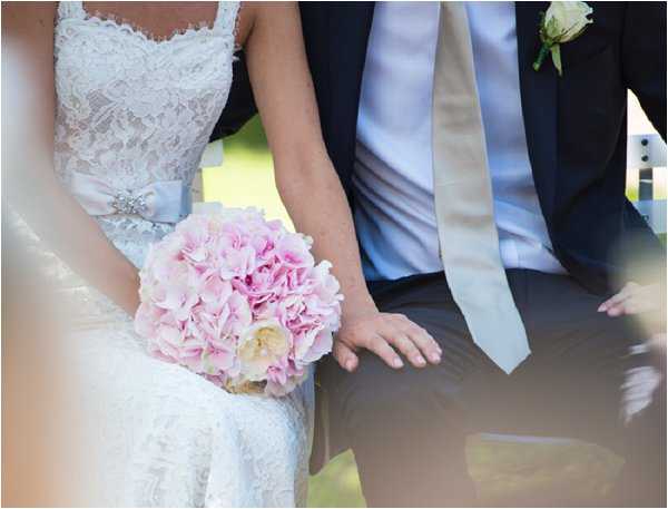 Close-up portrait of a bride and groom seated side by side, likely during an outdoor ceremony or portrait session. The bride wears a white lace dress with a pale blue satin sash accented by a rhinestone brooch, and holds a rounded bouquet of soft pink hydrangeas and cream garden roses. The groom is dressed in a navy suit with a light grey tie and a cream rose boutonniere. The bride's hand rests on the groom's leg, and the composition is tightly cropped to show only their torsos and hands.