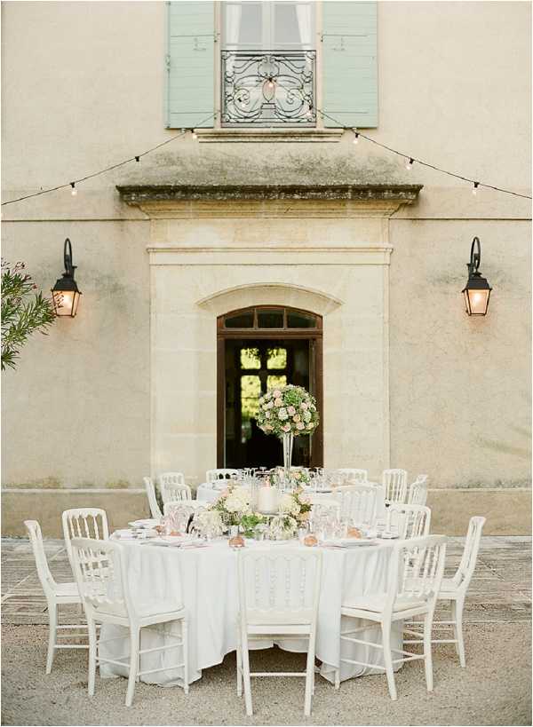 An outdoor reception table setup photographed against the facade of a French chateau, with no guests present. The round table is dressed in a white floor-length linen and surrounded by white Napoleon-style chairs. The centerpiece features a tall glass vase arrangement of blush pink and cream flowers, likely roses and hydrangeas, with greenery, while lower floral clusters in similar blush and ivory tones are also visible on the table. Place settings with glassware, pink napkins, and silver accents are arranged around the table. Two black wall-mounted lanterns flank a tall arched wooden door on the chateau's stone facade, and a string of bistro lights is strung above the table. The overall decor palette is white, blush, and soft gold with a classic French style. Wide shot framing the full table and building facade. Potential venue feature image.