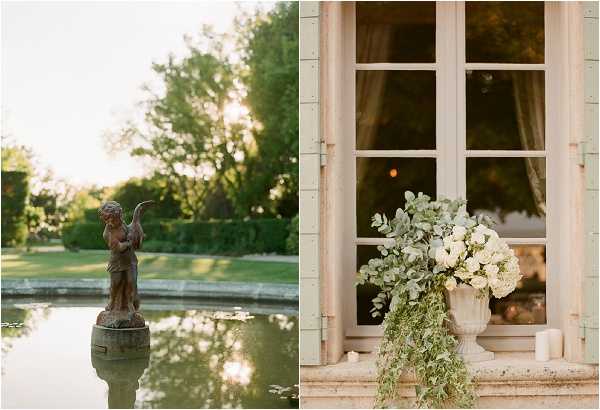 A split image showing two venue detail shots at what appears to be a French chateau property. The left frame features a classical stone cherub statue standing on a pedestal in the center of a formal reflecting pool, surrounded by manicured hedges and mature trees in warm evening light. The right frame is a close-up detail shot of a large white stone urn overflowing with white hydrangeas, garden roses, and cascading eucalyptus and trailing greenery, placed on a window ledge beside French doors with aged white shutters; two white pillar candles sit nearby, and warm candlelight is visible through the glass. The overall decor palette is white and green with a classic, formal French estate aesthetic. Potential venue feature image.