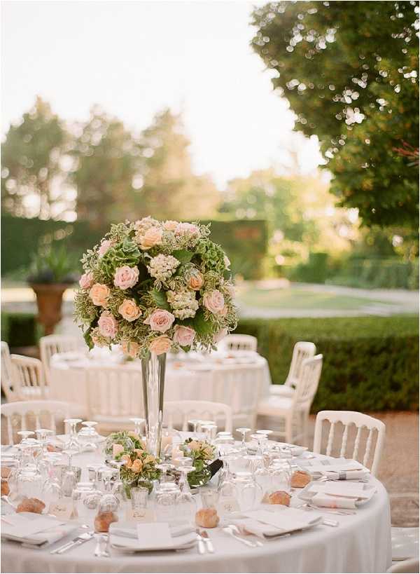 An outdoor wedding reception tablescape set in a formal garden, likely at a French chateau or estate. The round tables are dressed with white linens and set with white place settings, glassware, folded napkins, and small bread rolls at each place; white Napoléon-style chairs surround the tables. The focal point is a tall silver trumpet vase centerpiece holding a large spherical arrangement of blush and peach roses, green hydrangeas, and mixed greenery, with a smaller low floral cluster of matching blooms placed beside it on the table. The overall decor palette is white, blush, peach, and soft green, giving the setting a classic, formal French style. The shot is a medium close-up focused on the centerpiece with additional reception tables visible and softly blurred in the background.