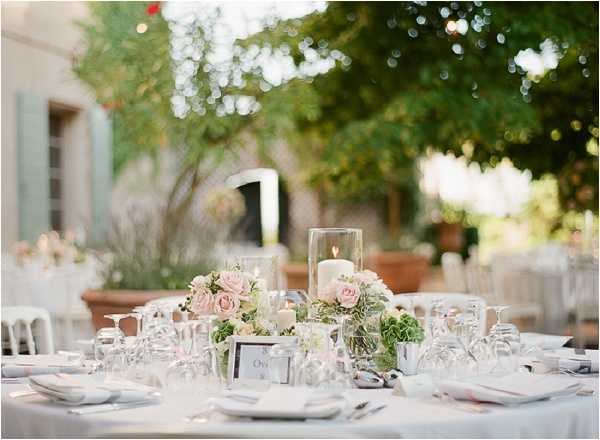 An outdoor wedding reception tablescape set in a courtyard, with a stone building featuring sage green shutters visible in the background. The round table is dressed in a white linen and set with white china, folded napkins, silver cutlery, and multiple wine glasses arranged around each place setting. The low centerpiece features blush pink roses and green hydrangeas arranged around a pillar candle in a glass hurricane vase, with small votive candles and a framed table number card completing the arrangement. The styling palette is soft white and blush pink with greenery accents, giving a classic romantic tone; the shot is a close-up detail image with shallow depth of field, with additional guest tables and large terracotta planters softly blurred in the background.