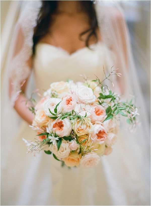 A close-up detail shot of a bride holding her bouquet, with the bride intentionally blurred in the background. The bouquet is a rounded arrangement composed of peach, blush, and cream garden roses and ranunculus, accented with small white filler flowers and trailing green foliage. The bride is wearing an ivory strapless sweetheart-neckline gown and a lace-edged veil that drapes over both shoulders. The overall floral palette is soft peach and cream with green accents, styled in a romantic, classic manner.