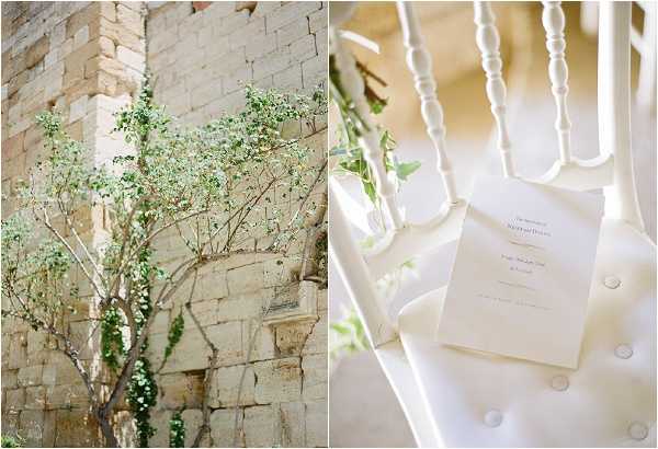 A split diptych image showing two detail shots from a wedding. The left panel features an outdoor stone wall with warm golden-toned ashlar masonry, with a small flowering tree and trailing green ivy growing against it. The right panel is a close-up detail shot of a white ceremony program booklet placed on the white cushioned seat of a white spindle-back chair, with a trail of green vine foliage draped across the chair back. The overall decor palette is all-white and green, suggesting a classic, clean aesthetic. Potential venue feature image.