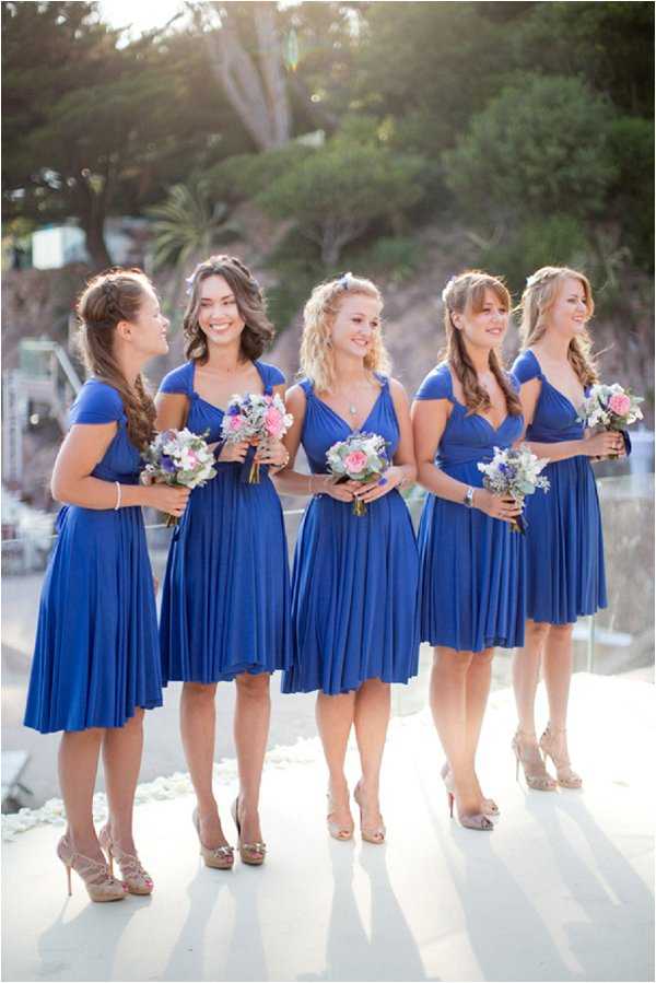 A bridal party portrait featuring five bridesmaids standing together outdoors on a white surface, likely a terrace or platform. All five women wear matching cobalt blue knee-length wrap-style dresses with cap sleeves and nude strappy heels. Each bridesmaid holds a small bouquet featuring pink roses, dusty blue flowers, and soft white blooms with silver-green foliage. The women wear their hair down or in soft half-up styles, and several have braided accents. The composition is a medium-wide portrait shot taken at golden hour with warm backlighting, with a softly blurred outdoor backdrop visible behind the group.