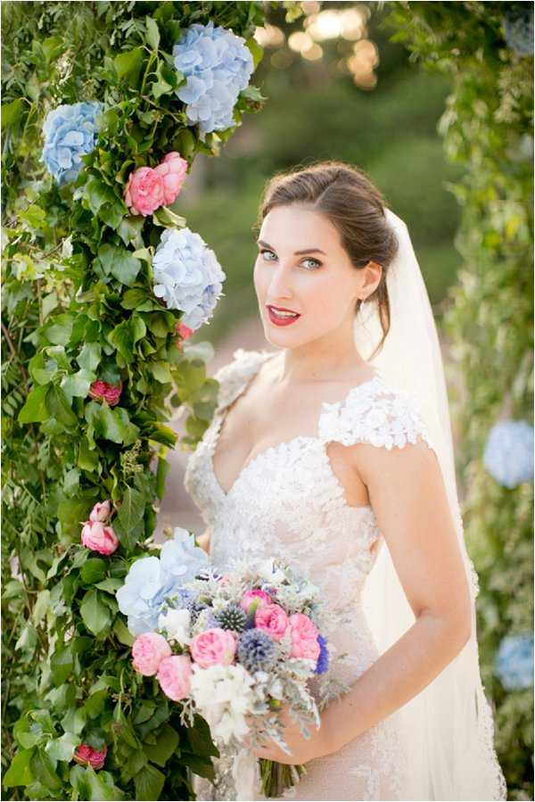 A bridal portrait taken outdoors, showing the bride standing beside a lush floral arch draped in green ivy and decorated with powder blue hydrangeas and pink garden roses. The bride wears an ivory lace gown with lace cap sleeves and a flowing veil, and has her dark hair swept up; she wears a bold red lip. She holds a mixed bouquet featuring pink garden roses, blue globe thistles, white blooms, lavender, and silver-toned foliage. The color palette of the florals combines powder blue, hot pink, white, and purple tones. The shot is a medium portrait with a softly blurred outdoor background.
