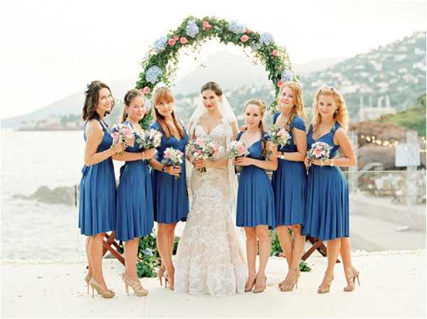 A bridal party portrait taken outdoors at a coastal venue, showing a bride flanked by six bridesmaids posing in front of a circular floral arch. The bride wears a fitted lace gown with a sweetheart neckline and a cathedral veil, holding a bouquet of pink and white blooms. The six bridesmaids wear matching cobalt blue knee-length wrap-style dresses with nude heeled sandals, each carrying bouquets of pink and white flowers including what appear to be hydrangeas and roses. The circular arch behind the group is decorated with lush greenery, blue hydrangeas, and pink roses. The setting appears to be a waterfront terrace or ceremony platform with the sea and hillside visible in the background. The image is a wide group portrait shot with a soft, film-like quality.