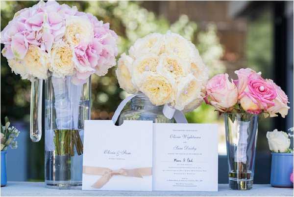 A detail flat-lay style shot of a wedding stationery and floral display arranged on a table, photographed outdoors in natural light. Three glass vases hold distinct floral arrangements: a bouquet of soft pink hydrangeas and cream garden roses on the left, a tightly packed ivory garden rose bouquet in the center, and a smaller arrangement of pink standard roses and pink ranunculus on the right. In the foreground, two white wedding stationery pieces are propped up — a folded program tied with a blush satin ribbon and an open wedding invitation card reading the names 'Olivia Wightman' and 'Sean Darby' in black script. The decor palette is soft and romantic, featuring blush, ivory, and pink tones against a clean white and pale blue background.