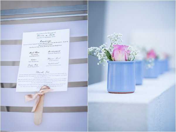 A split detail shot featuring two close-up images of wedding decor elements. On the left, a printed ceremony program printed on white card is tied with a small blush satin ribbon bow, listing readings, the best man, maid of honor, ushers, and officiant along with a thank-you message. On the right, a row of small periwinkle blue ceramic mugs or pots, each containing a single pink rose paired with white baby's breath, are lined up along a white surface, receding into a softly blurred background. The overall decor palette combines periwinkle blue, blush pink, and white in a simple, classic style.