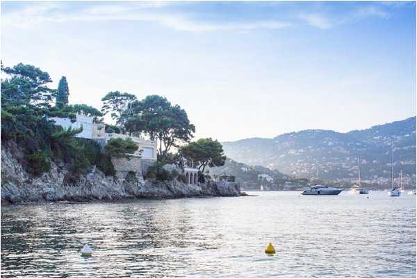 Wide shot taken from the water looking toward a white villa perched on a rocky coastal cliff, surrounded by pine trees, with a calm bay in the foreground where several yachts and sailboats are moored. Rolling hills dotted with buildings extend across the background in soft haze. No people or wedding elements are visible in this image. Potential venue feature image.