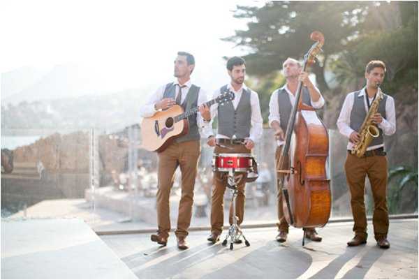 A four-piece live band performs at an outdoor wedding, positioned on a terrace or paved platform with a coastal or hillside backdrop visible in the soft, hazy background light. The musicians — playing acoustic guitar, snare drum, double bass, and saxophone — are dressed in a coordinated style of white dress shirts, grey vests, brown trousers, and leather shoes. The wide shot captures all four performers standing in a loose line, with natural backlighting creating a bright, washed-out atmosphere. The overall styling is classic-casual with a vintage jazz band aesthetic.