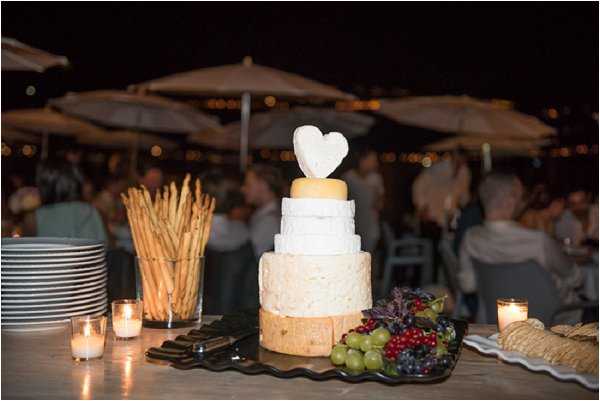 A detail shot of a wedding cheese tower display at an outdoor evening reception, styled to resemble a tiered cake. The tower is composed of four stacked whole cheese wheels of varying sizes, topped with a heart-shaped piece of cheese. The display table includes breadsticks in a tall container, a platter of green and red grapes alongside dark berries, stacked white plates, chocolate bars, crackers, and small lit votive candles. In the blurred background, a crowd of wedding guests mingles under large cream patio umbrellas with warm string lights visible in the distance, suggesting a terrace or outdoor venue setting at night.