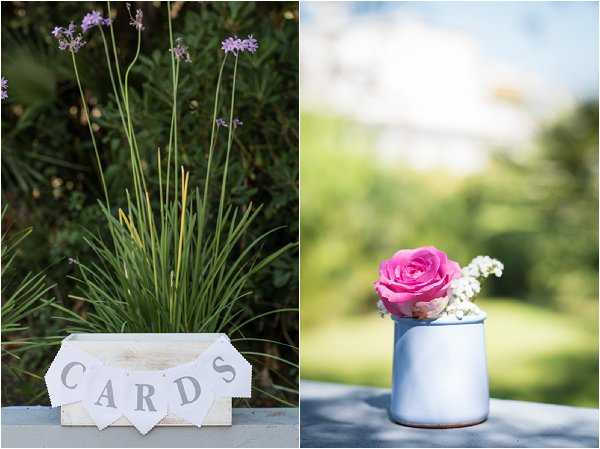 A two-image detail collage showing rustic wedding decor shot outdoors. On the left, a whitewashed wooden card box with a white pennant banner spelling 'CARDS' sits on a surface in front of naturally growing lavender stalks. On the right, a small white enamel milk jug holds a single bright pink rose alongside sprigs of white baby's breath. Both shots are close-up detail compositions with soft background blur, consistent with a rustic or boho decor style using a palette of white, grey-blue, and pink accents.