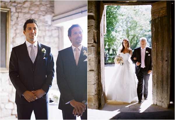 A diptych of two ceremony moments. On the left, the groom and a groomsman stand waiting inside a venue, both wearing dark navy suits with light pink ties and white boutonnières, hands clasped in front of them. On the right, the bride is being walked down the aisle by an older man, likely her father, who wears a grey morning suit with a pink tie; they are entering through a large stone archway doorway with bright natural light behind them. The bride wears a strapless white ballgown and carries a loose, rounded bouquet of ivory and blush flowers with greenery. The setting appears to be a rustic stone chapel or historic venue in France. Both shots are medium portrait-style compositions with natural daylight.