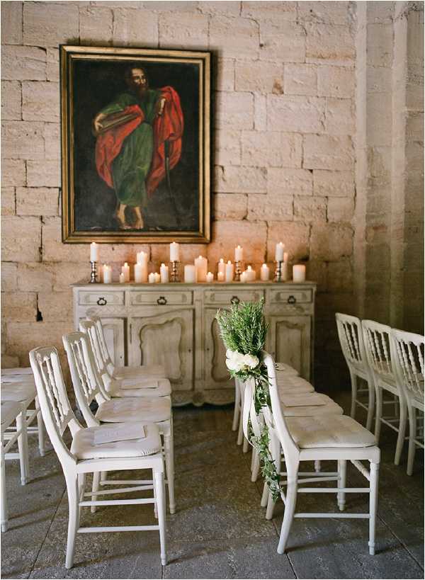 An indoor wedding ceremony setup inside a historic stone-walled venue, likely a chapel or chateau. Rows of white painted wooden chairs are arranged on either side of a central aisle, with the end chair decorated with white blooms and a trailing greenery garland. A distressed grey French provincial sideboard serves as a backdrop, topped with an arrangement of white pillar candles and taper candles in brass holders creating warm ambient lighting. Above the sideboard hangs a large oil painting in a gilt frame depicting a robed religious figure. The decor palette is neutral and muted — white chairs, soft candlelight, and green-and-white florals — with a classic, understated French interior style. Wide shot capturing the full ceremony space. Potential venue feature image.