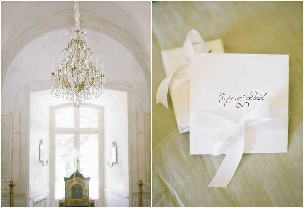 A two-image detail flat lay composite. On the left, a wide interior shot captures a ornate gold and crystal chandelier hanging beneath a arched ceiling in a formal venue, with tall arched windows and wall sconces visible in the background and a painted antique chair in the foreground, suggesting a chateau or palace setting. On the right, a close-up detail shot shows an ivory square wedding ceremony booklet or invitation tied with a wide ivory satin ribbon bow, placed on a soft sage-green linen surface; the booklet features calligraphy script reading what appears to be the couple's names. The overall palette is ivory, cream, and soft gold, with a classic French chateau aesthetic.