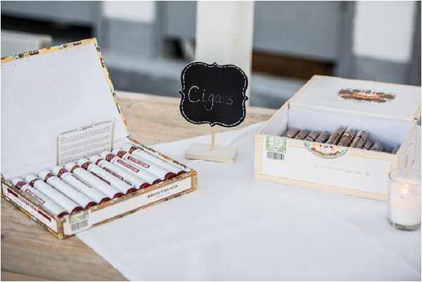 A close-up detail shot of a cigar station set up at a wedding reception. Two open cigar boxes are displayed on a white linen-covered wooden table — one containing individually wrapped cigars in white tubes with dark red bands, and another with unwrapped cigars. A small chalkboard sign on a wooden stand reads 'Cigars' in handwritten chalk lettering. A small votive candle in a glass holder is visible at the right edge of the frame. The styling is clean and rustic, consistent with a relaxed reception atmosphere.