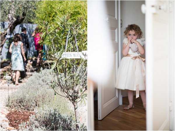 A diptych of two wedding images. Left image: an outdoor garden or grounds scene where a small group of guests walks along a path lined with olive trees and lavender plants, following a small white directional sign reading 'THIS WAY'; guests are dressed in casual summer attire including a light blue floral dress. Right image: a close-up portrait of a young flower girl, approximately toddler age, standing in a doorway; she wears an ivory tulle dress with a champagne/gold satin sash and has curly hair, with her hands raised near her face in a shy or curious pose.