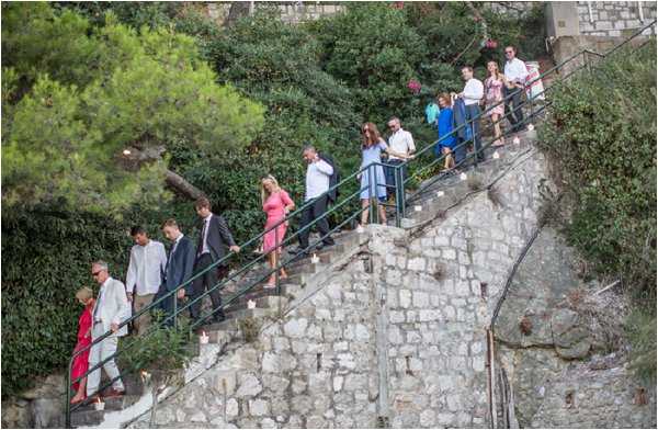 A group of approximately 15 wedding guests descends a wide stone staircase with metal railings at an outdoor venue, likely a coastal or hillside property in the south of France or similar Mediterranean setting. The guests are dressed in smart-casual to formal attire, including a coral-pink dress, a red dress, a light blue dress, and dark suits. Small white votive candles are placed along the base of the stone steps as decorative lighting elements. The wide shot captures the full length of the staircase framed by dense green foliage and a stone retaining wall.