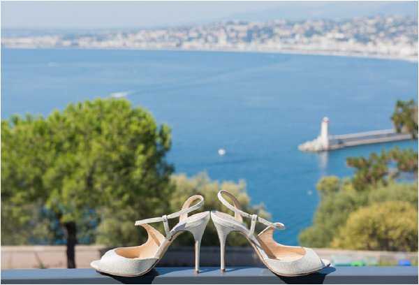 A detail close-up shot of a pair of ivory glitter slingback stiletto heels placed on a railing or ledge, with a sweeping Mediterranean coastal view in the soft-focus background, including a harbor with a lighthouse and a town along the shoreline. The shoes appear to be open-toe with ankle straps and a high stiletto heel, likely bridal footwear. The composition frames the shoes as the sharp focal point against the blurred blue water and coastal scenery behind them.