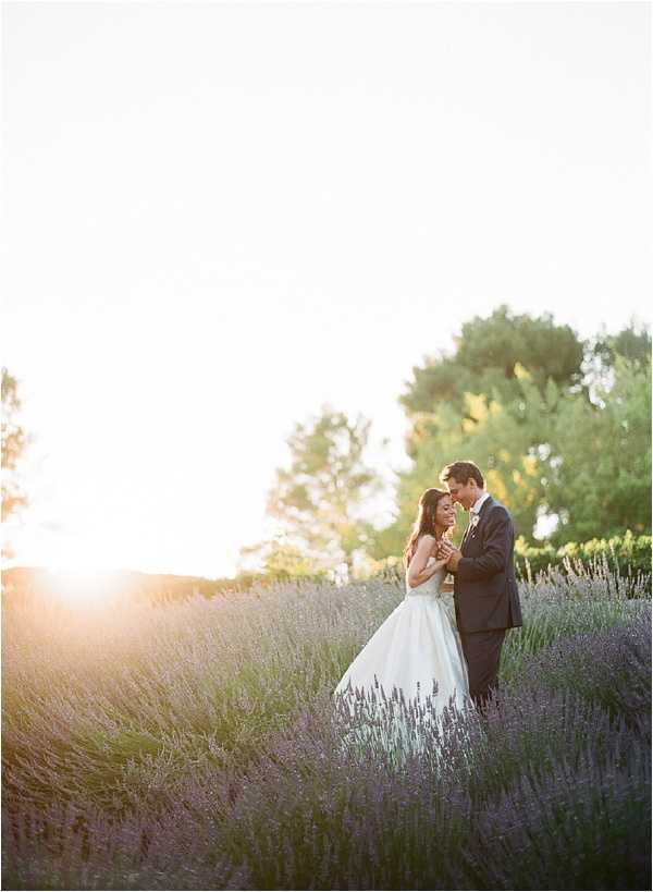 A couple portrait taken outdoors in a lavender field at golden hour, with warm backlight creating a strong sun flare from the left side of the frame. The bride wears a white ball gown with a fitted bodice and full skirt, while the groom is dressed in a dark navy suit; the two stand close together, foreheads nearly touching, holding hands. Rows of purple lavender extend across the mid-ground, and mature green trees are visible in the background. The composition is a medium full-length portrait shot with the couple positioned to the right of center.