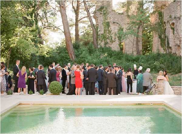 A cocktail hour or reception gathering of approximately 40-50 guests mingling poolside at an outdoor venue featuring stone ruins and mature trees in the background. The pool in the foreground has pale turquoise water, and guests are standing on a light stone terrace along its edge. The bride, visible on the far right, wears a white gown, while guests are dressed in a mix of formal attire including dark suits, a red dress, a purple dress, and blush pink dresses. The setting has a rustic, historic character with ivy-covered stone walls and ancient ruins forming a dramatic backdrop. Wide establishing shot taken from across the pool.