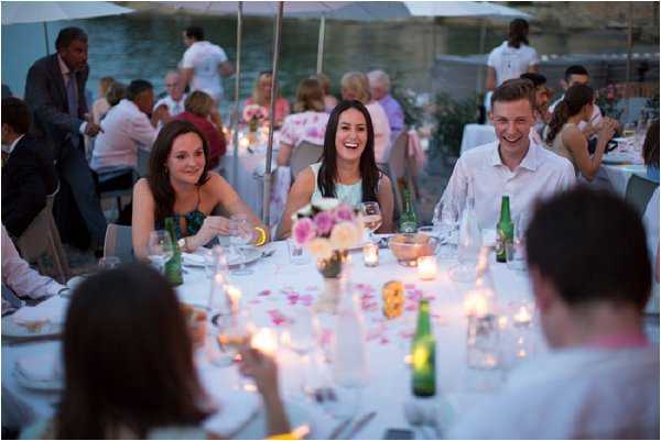 Wedding reception dinner scene shot outdoors in the early evening, with guests seated at a round white-linen table laughing and socializing. The foreground shows two women and a man in animated conversation — one woman wearing a teal/green dress, another in a white top, and the man in a white dress shirt. The table is decorated with small clusters of pink and white flowers, scattered rose petals, votive candles, and green glass beer bottles alongside wine glasses and water carafes. The warm candlelight creates a soft glow across the table in the dimming light. Additional guests are visible at surrounding tables in the background. The composition is a medium candid shot taken from a low angle at table level.