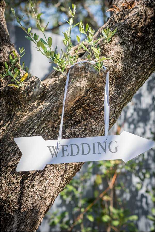 A close-up detail shot of a white arrow-shaped directional sign reading 'WEDDING' in grey lettering, hung from a tree branch using white ribbon. The sign points to the right and is suspended in the fork of a textured tree trunk with small leafy branches growing nearby. The styling is classic and simple, using a white metal or painted wooden arrow sign as wayfinding decor for guests.