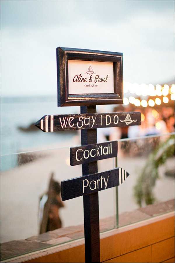 Close-up detail shot of a decorative wedding directional sign post positioned outdoors at a beachside venue during dusk. The dark-stained wooden sign features a framed panel at the top reading 'Alina & Pavel' with a small sailboat illustration, and two arrow-shaped directional boards below reading 'We Say I Do' and 'Cocktail Party,' also decorated with sailboat motifs and striped details. The styling follows a nautical theme with dark wood and cream lettering. In the soft-focus background, warm string lights and what appears to be a set reception area are visible, with a sandy beach and water beyond a glass barrier.