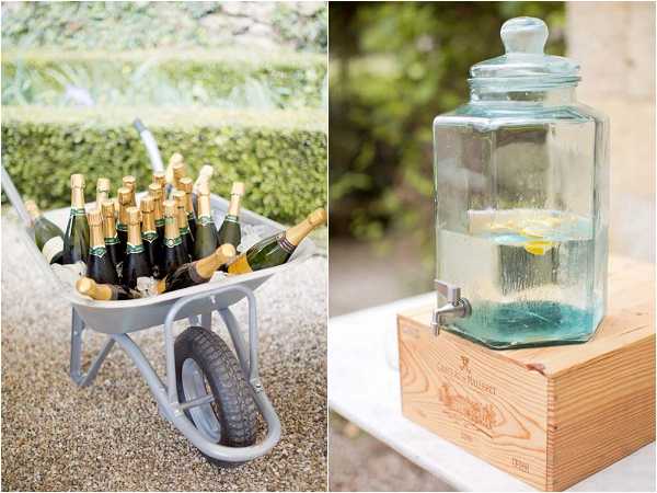 A split-image detail shot of outdoor cocktail hour drink stations at a garden wedding. On the left, a grey metal wheelbarrow is filled with ice and approximately 15 bottles of champagne with gold foil tops, styled as a rustic drinks display on a gravel surface. On the right, a large glass beverage dispenser with a silver spigot holds water with lemon slices and is elevated on a natural pine wood wine crate, adding a rustic, countryside styling touch. Both images are bright and airy with soft natural lighting.