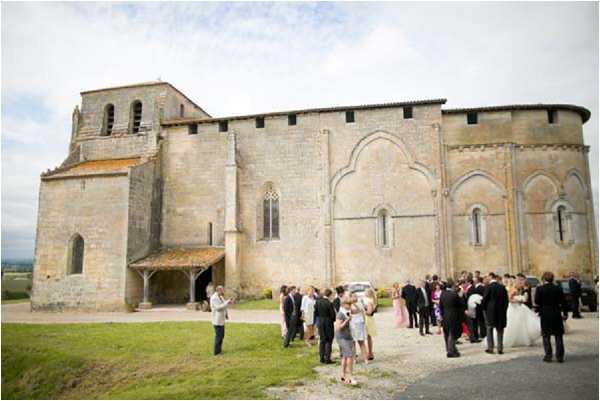 Wedding guests gather outside a Romanesque stone church following what appears to be a ceremony, with approximately 30–40 people mingling on the gravel forecourt. The church features characteristic rounded apse architecture, arched blind arcading, and a small bell tower, typical of medieval French religious buildings. The bride is visible in a full white ballgown and the guests are dressed in formal attire, including dark suits and colourful occasion dresses. The image is a wide exterior shot taken from a slight distance, capturing both the full scale of the church building and the crowd below. Potential venue feature image.