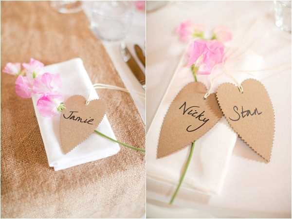 Close-up detail shot of wedding place settings featuring kraft paper heart-shaped name cards with scalloped edges, handwritten with guest names 'Jamie,' 'Nicky,' and 'Stan' in black ink. Each place setting includes a single stem of pink sweet peas tucked alongside a white folded napkin, with the name card tied on using twine. The left setting rests on a burlap/hessian table runner over a warm gold-toned tablecloth, while the right shows a white linen setting. The styling is rustic with a warm neutral and soft pink color palette.
