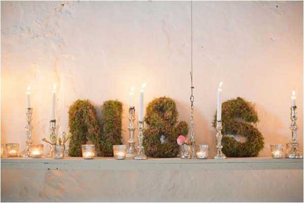 Close-up detail shot of a wedding reception decor display arranged on a narrow shelf or mantle against a textured off-white plaster wall. The centerpiece features three large moss-covered letters spelling 'N & S,' serving as monogram initials for the couple, with a small pink floral bloom tucked into the ampersand. The display is flanked and interspersed with silver candelabras holding tall white taper candles and small glass votive holders with lit tea light candles, casting a warm amber glow. The overall styling is rustic-organic with natural green moss letters paired with metallic silver candleholders, suggesting a soft, nature-inspired decor palette.
