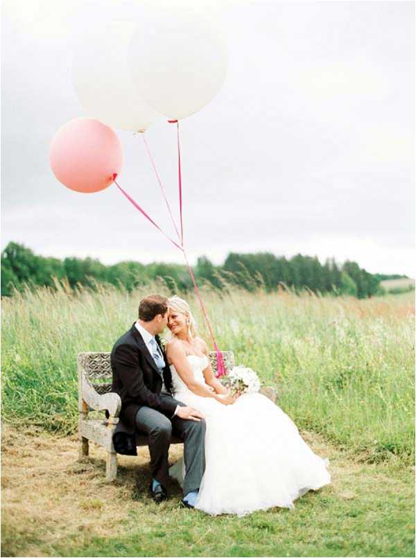A couple portrait taken outdoors in an open meadow setting, with the bride and groom seated together on a weathered stone or concrete bench. The groom wears a dark navy suit with a light blue tie, and leans in toward the bride, who is dressed in a strapless white ballgown and holds a small bouquet of white flowers with greenery. Three oversized round balloons — one white and two in coral/blush pink — are tied with bright pink ribbon and held by the couple, floating above them. The styling is relaxed and playful with a soft pastel color palette. Medium-distance portrait shot with the couple centered in the frame against a backdrop of tall meadow grasses.
